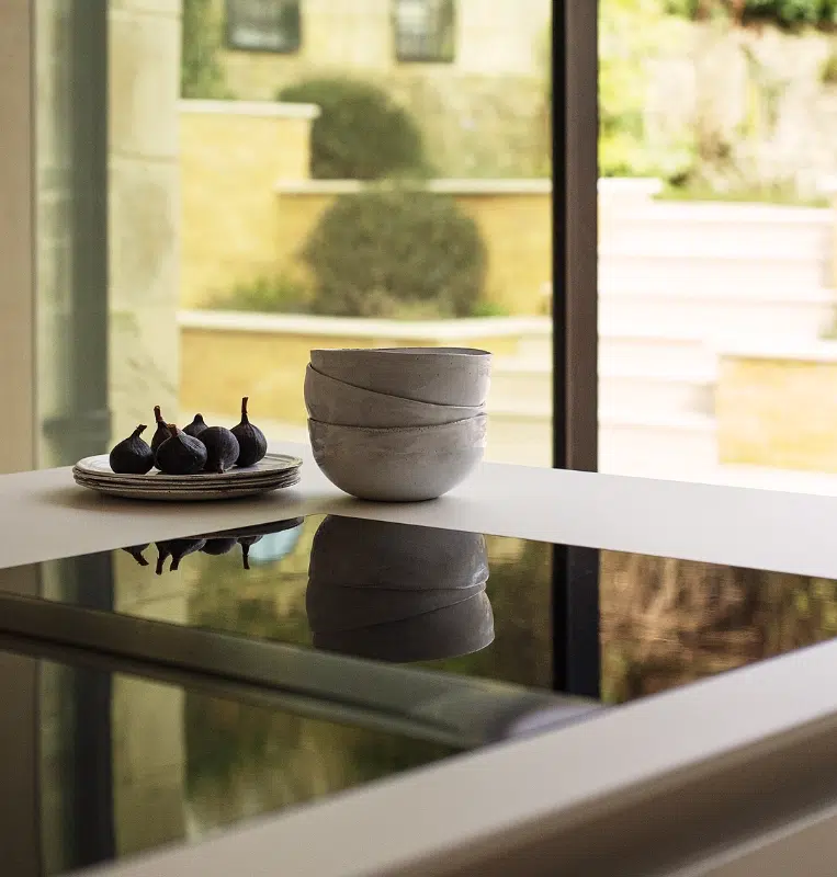 Black induction hob set into a reflective worktop beside a window overlooking clipped garden topiary.