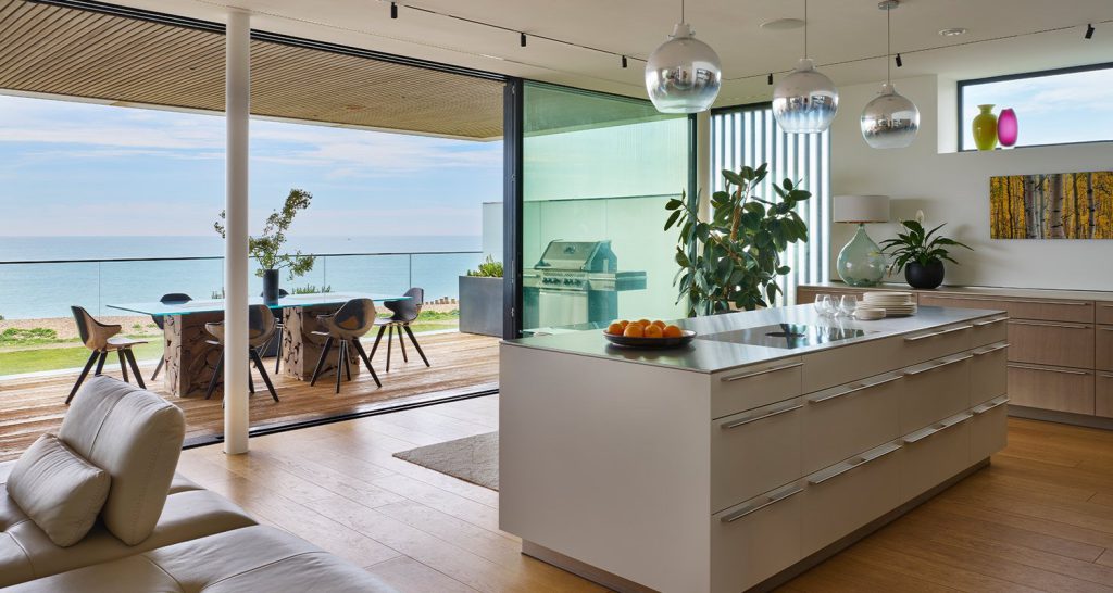 White kitchen island beside full-height glazing overlooking the living area and sea