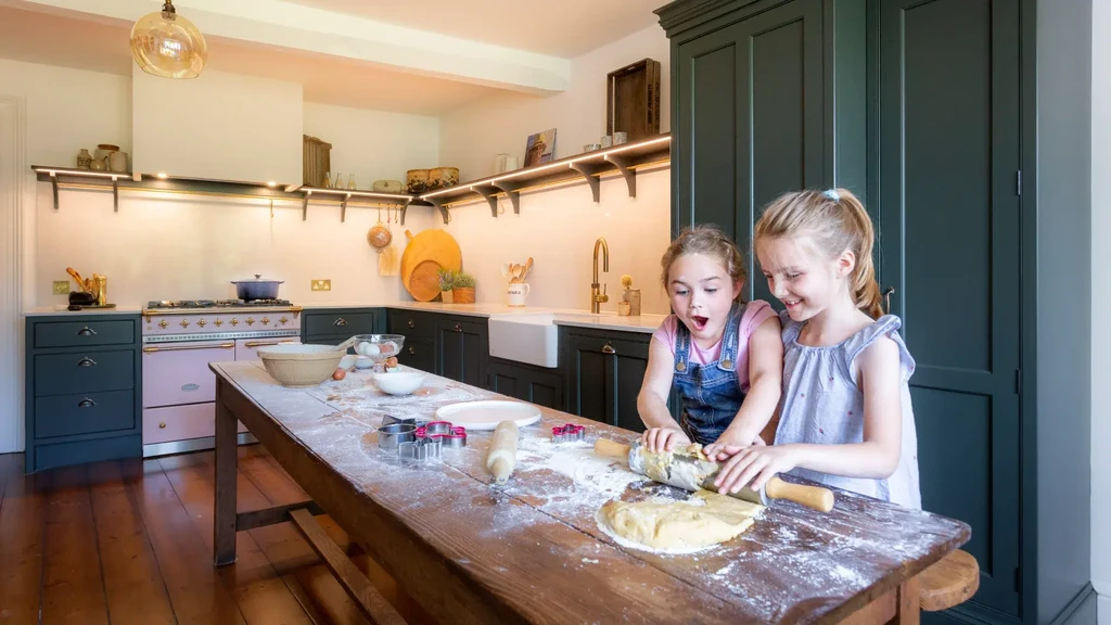 Two children baking at a timber table in a painted Craftsman Collection kitchen with open shelving and dark green cabinetry.
