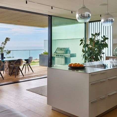 White kitchen island beside full-height glazing, with views across the adjoining living area towards the sea.