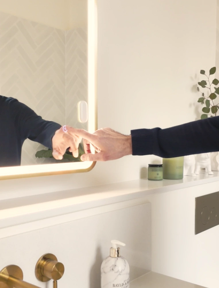 A hand reaching beneath the illuminated bathroom mirror, showing the accessible position above the vanity.