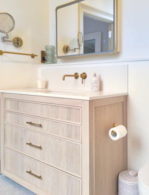 Pale fluted vanity with quartz top, wall-mounted brass tap, illuminated mirror and brass toilet-roll holder.