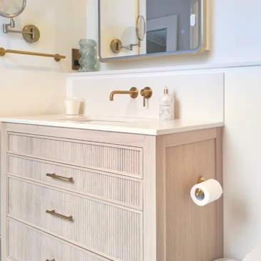 Pale fluted vanity with quartz top, wall-mounted brass tap, illuminated mirror and brass toilet-roll holder.