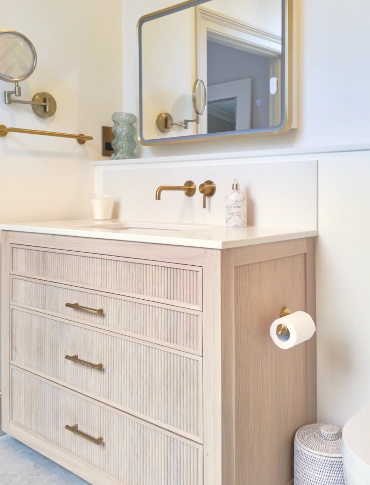 Pale fluted vanity with quartz top, wall-mounted brass tap, illuminated mirror and brass toilet-roll holder.