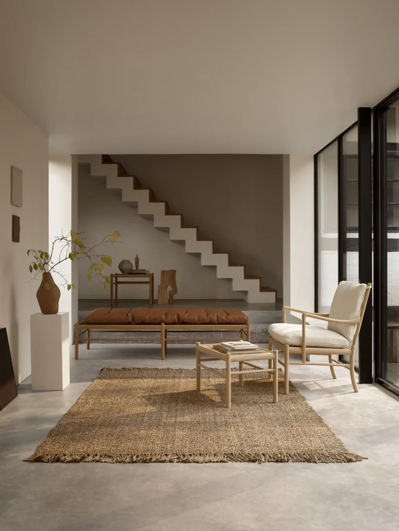 Timber-framed lounge chair and footstool in a neutral living room beneath an open staircase, with a woven rug and low table.